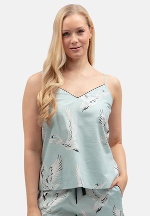 Vrouw met lang blond haar die een lichtblauwe cami en korte broek met witte zwaanprints draagt, staat met één hand in de zak.