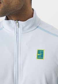 Gaiši zils jaka ar augstu apkakli, ar rāvējslēdzēju un zaļu Nike logotipu dzeltenā apmalē kreisajā krūtī. Gluda tekstūra.
