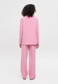 Roze op maat gemaakt pak met een lange blazer en wijde broekspijpen, vervaardigd uit soepel materiaal, met een subtiele rugnaad en een minimalistisch ontwerp.