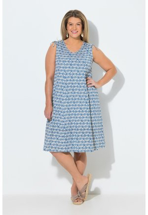 Kvinde smilende, iført en blå og hvid mønstret ærmeløs knælang kjole med bundne skulderstropper og beige kilehælssandaler.