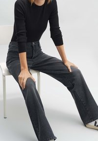 Personne assise sur une chaise blanche portant un haut à manches longues noir, un jean large taille haute gris foncé et des baskets blanches avec des accents noirs.