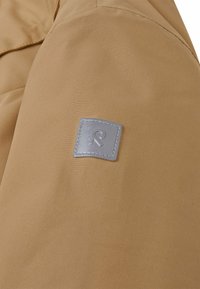Brauner, strukturierter Stoff mit einem silbernen rechteckigen Logo-Patch, das ein stilisiertes "R" mit Nähdetail auf einer Jacke zeigt.