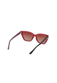 Gafas de sol de gran tamaño en forma de ojo de gato, rojas, con lentes en degradado. Las varillas negras tienen un patrón geométrico rosa. Montura de plástico ligera.