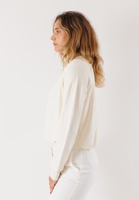 Donna con capelli castano chiaro mossi, che indossa un maglione bianco a maniche lunghe e pantaloni bianchi, in piedi di profilo su uno sfondo semplice.
