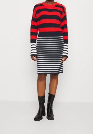 Personne portant une robe en maille rayée rouge et marine avec des rayures blanches sur les manches et la jupe, associée à des bottes noires à talons mi-mollet.