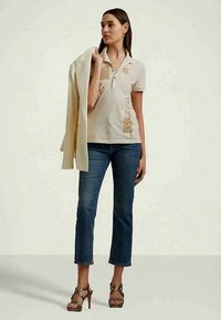 Sieviete, kas ģērbusies bēšā polo krekliņā ar ziedu motīviem, zils īsās bikses, brūnās augstpapēžu sandales, tur vieglu jaku pār plecu, stāvot pret vienkāršu fona.