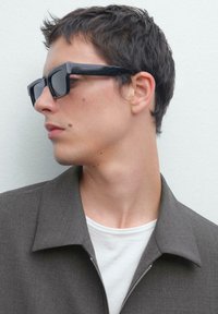 Sorte rektangulære solbriller med mørke linser, båret med en grå zip-up jakke over en hvid skjorte, der fremhæver rene linjer og moderne design.