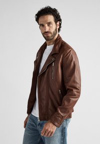 Brun lädermotorcykeljacka med strukturerad yta, asymmetrisk dragkedjestängning och två dragkedjefickor. Vit t-shirt och ljusblå jeans under.