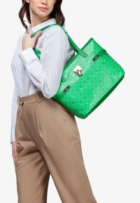 Borsa verde realizzata in materiale lucido, con un motivo geometrico, doppi manici e una chiusura metallica.