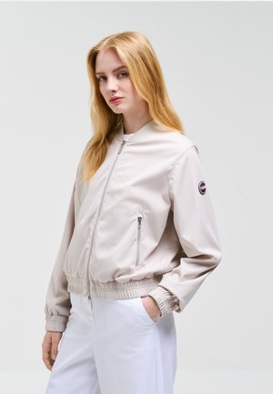 Jeune femme aux longs cheveux blonds portant une veste bomber beige clair et un pantalon blanc, posant avec une main dans la poche devant un fond uni.