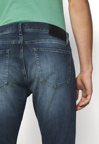 Nahaufnahme einer Person, die ausgewaschene blaue Jeans und ein grünes Hemd trägt, wobei die Gesäßtaschen und der rechte Arm seitlich sichtbar sind.