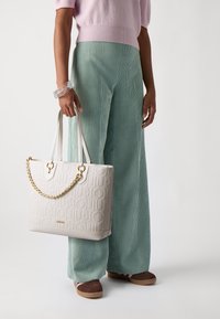 Borsa a tote bianca con trama e accentuazione a catena, caratterizzata da un design in rilievo del logo. Il modello indossa pantaloni di velluto a coste verde chiaro e una maglietta rosa.
