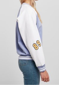 Světlomodrá a bílá mikina se sportovním designem, s výšivkou "89" na rukávu a žebrovanými manžetami a lemem.