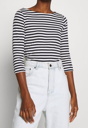 Persoon die een zwart-wit horizontaal gestreept shirt met lange mouwen draagt, ingestopt in een high-waisted lichtblauwe spijkerbroek, tegen een effen achtergrond.