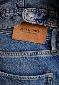Kerged teksad helesinises toonis, millel on kulunud tekstuur. Tuhkbež värvi nahapatch, millel on musta värvi kiri "JACK & JONES", asub vöökoha juures.
