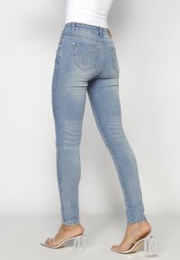 Jeans skinny de denim azul claro con un diseño curvado en los bolsillos traseros, realzados con costuras decorativas y sandalias de tacón de plástico transparente.