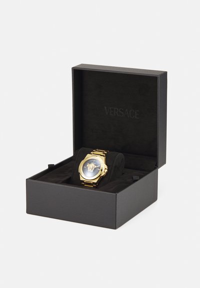 Zelts pulkstenis ar zilu ciparnīcu un Medūzas logotipu, ievietots melnā Versace kastē, kas izklāta ar melnu samtu. Kastē nav papildu detaļu.
