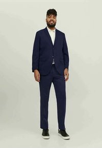 Man with beard wearing navy blue suit, white shirt, and black sneakers standing against plain light background.