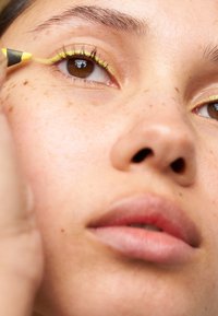 Grønn og gul eyeliner på brune øyne, med synlige fregner og jevn hudtekstur. Myke rosa lepper og naturlig sminke.