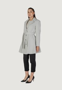 Trench coat grigio chiaro con finitura testurizzata, bottoni doppi e cintura, abbinato a pantaloni neri a tre quarti e tacchi.