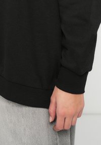 Το χέρι χαλαρό στο πλάι, φορώντας μια μαύρη μακρυμάνικη μπλούζα με ριχτούς καρπούς και ανοιχτό γκρίζο παντελόνι, μπροστά σε απλό φόντο.