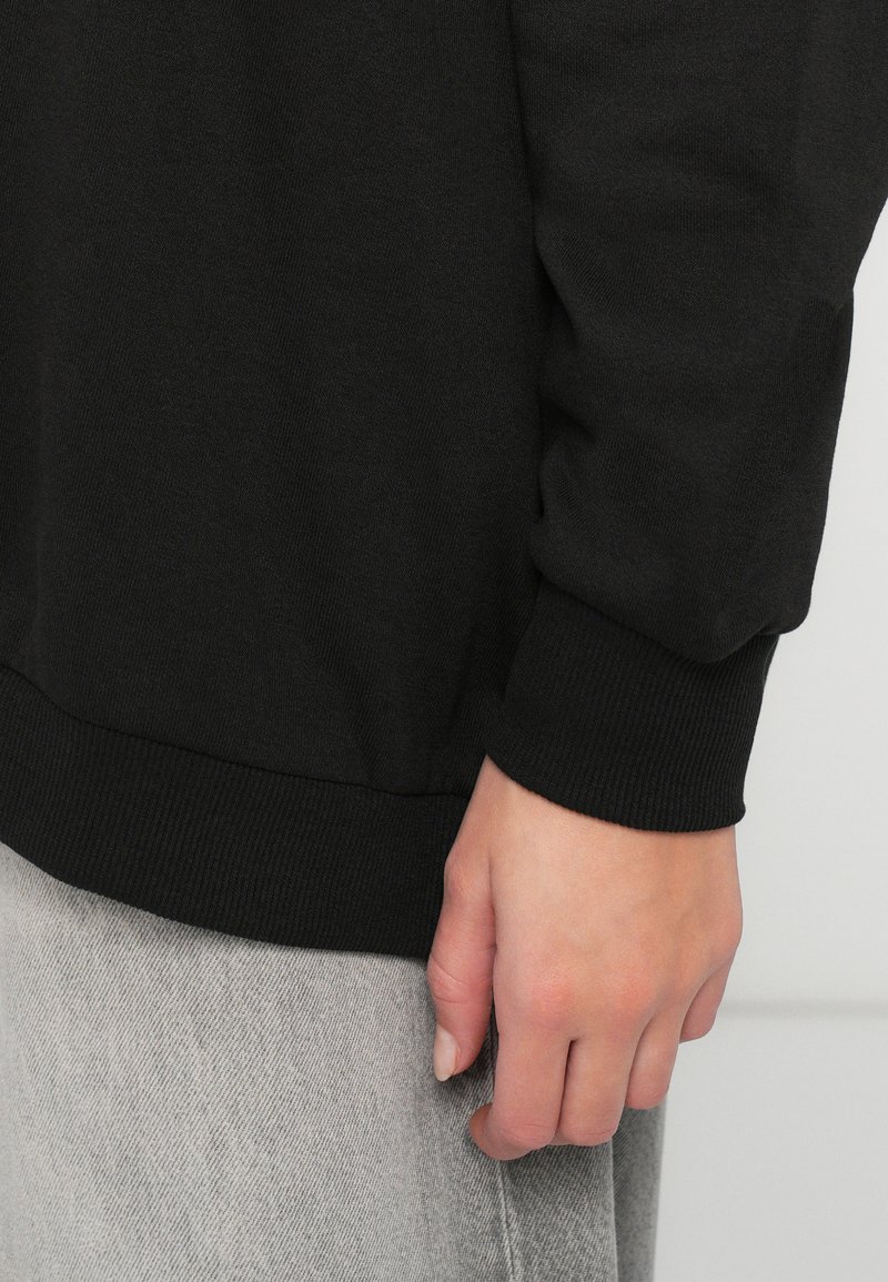 Hand ontspannen aan de zijkant, gekleed in een zwart overhemd met lange mouwen en geribbelde manchetten, en lichtgrijze broek tegen een effen achtergrond.