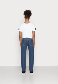 Camiseta blanca de manga corta con rayas negras en las mangas, combinada con jeans rectos de color azul oscuro con bolsillos traseros y una etiqueta marrón.