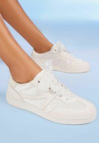 Vita sneakers gjorda av strukturerat läder och mesh, med ett prickigt mönster och sidenband. Sulan är platt och rund.