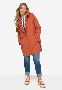 Oranssi vettä hylkivä takki, jossa on huppu, kaksi etutaskua ja napitus; yhdistettyinä käärityihin sinisiin farkkuihin ja oransseihin lenkkareihin.