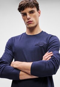 Mornarski dolg rokav s krožnim izrezom in teksturiranim logotipom na prsih. Mehak material z dolgimi rokavi, ki segajo do zapestij.
