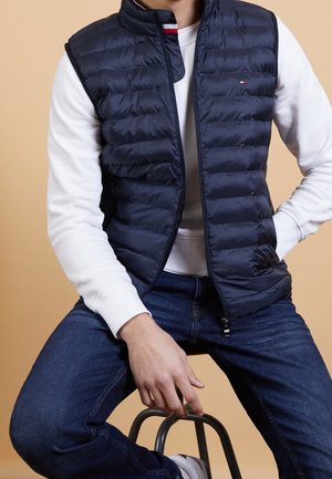 Middengrijze puffer vest met een quiltpatroon, hoge kraag en rits. Gedragen over een wit shirt met lange mouwen, gecombineerd met donkere spijkerbroek. Eenvoudige metalen accenten.