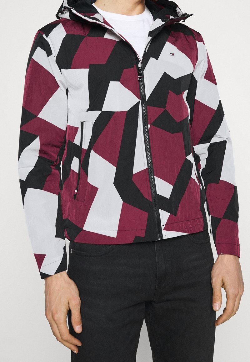 Homme portant une veste à capuche avec un motif géométrique marron, noir et blanc, assortie à un pantalon noir et une chemise blanche en dessous.