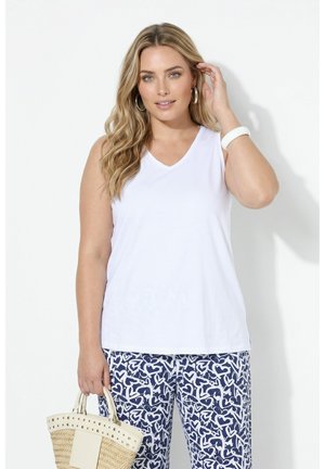 Femme aux longs cheveux blonds portant un haut blanc sans manches et un pantalon bleu à motifs, tenant un sac à main tissé beige.