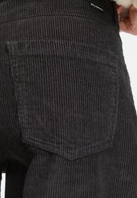Pantalon en velours côtelé noir avec une poche arrière à texture nervurée verticale et des coutures simples ; marquage minimal visible au niveau de la taille.