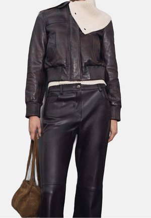 Osoba ubrana w ciemnobrązową skórzaną kurtkę i spodnie, kremowy dziergany sweter i szalik, trzymająca brązową zamszową torebkę przy boku.