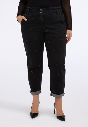 Kobieta nosząca czarne dżinsy z wysokim stanem i podwiniętymi nogawkami, z delikatnymi, błyszczącymi kwiatowymi detalami oraz czarne czółenka na wysokim obcasie z ostrym noskiem.