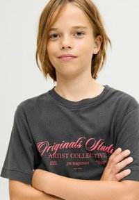 Joven con cabello castaño claro de pie con los brazos cruzados, lleva una camiseta gris oscuro con el texto rosa "Originals Studio Artist Collective."