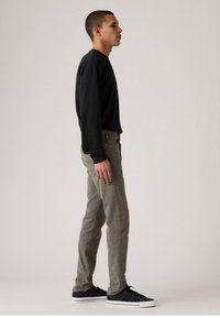 Black long-sleeve shirt, slim-fit olive green jeans, black sneakers with white soles; model standing sideways against a neutral background.