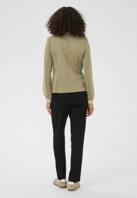 Light olive green long-sleeve top with a fitted design and white-striped cuffs, paired with black trousers and beige shoes. Smooth texture.