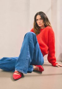 Roter, flauschiger Pullover, übergroße blaue Jeans und rote spitze Ballerinas mit Riemchen, die gegen einen neutralen Hintergrund sitzen.