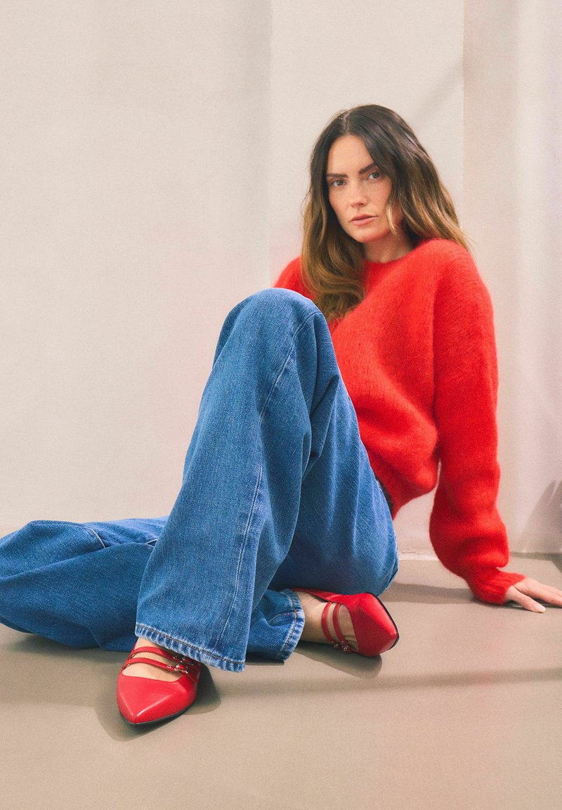 Roter, flauschiger Pullover, übergroße blaue Jeans und rote spitze Ballerinas mit Riemchen, die gegen einen neutralen Hintergrund sitzen.