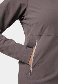 Chaqueta softshell gris con mangas largas, que presenta un bolsillo con cremallera y una textura suave. El diseño incluye costuras y un ajuste atlético.