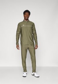 Oljčno zelena trenirka s zipped jakno in hlačami. Ima bele detajle in logotip Under Armour. Narejena je iz gladkega, elastičnega materiala.