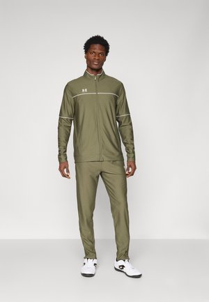 Maslinasto zeleni trenirka s jaknom na patentni zatvarač i hlačama. Sadrži bijele naglaske i Under Armour logo. Izrađena od glatkog, rastezljivog materijala.