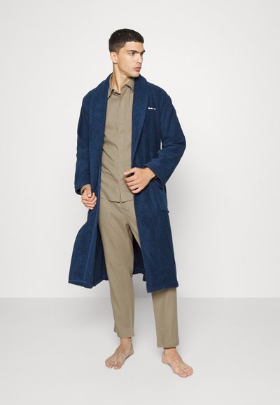 Robe de tecido felpudo azul-marinho com colarinho xale, bolsos laterais e design de comprimento médio, usado sobre calças leves bejes.