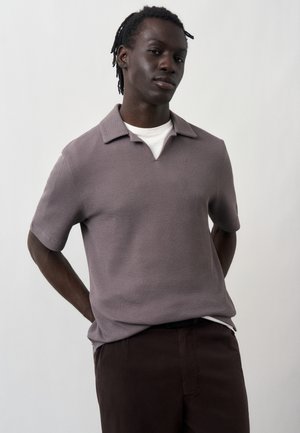 Νέος άνδρας με σκούρο δέρμα και κοντά dreadlocks που φοράει ένα πουκάμισο σε μωβ απόχρωση με κοντά μανίκια και γιακά πάνω από ένα λευκό t-shirt, στέκεται με τα χέρια πίσω από την πλάτη.