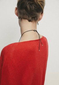 Pull en maille rouge à coupe ample. Un cordon noir orné de petites perles est visible à la nuque, mettant en valeur le détail du dos du vêtement.