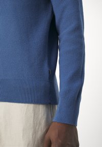 Gros plan d'une personne portant un pull bleu texturé avec des poignets côtelés et un pantalon beige, mettant l'accent sur la zone du bras et du torse.