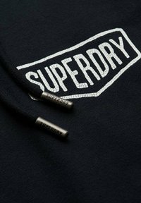Zwarte stof met een wit gebarsten Superdry-logo en twee zwarte trekkoorden met metalen uiteinden waarop "Superdry" gegraveerd staat.