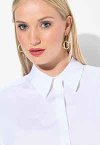 Witte hemd met knoopsluiting en een puntige kraag, gladde textuur en subtiele glans; model draagt grote gouden creolen.